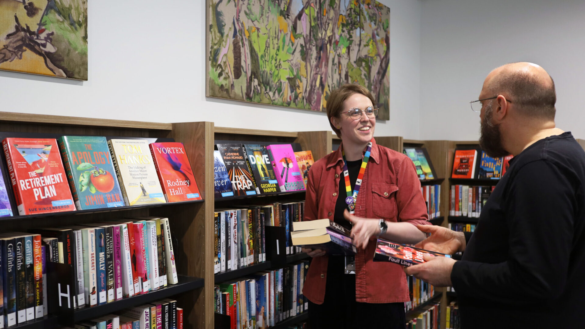 Two people stand in front of a shelf of books in Ascot Vale Library. They are both holding books. One of them has a rainbow staff lanyard on.